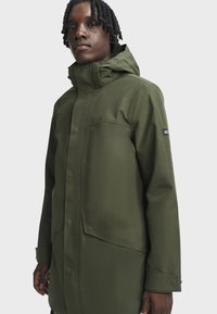 Olivově zelená nepromokavá bunda s kapucí, patentními uzávěry, velkými předními kapsami a strukturovaným povrchem. Obsahuje logo na rukávu.