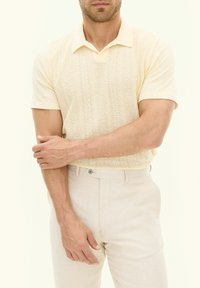 Stickad gul polotröja med korta ärmar, prydd med ett strukturerat diamantmönster, tillsammans med ljusbeiga strukturerade byxor.