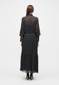 Γυναίκα που στέκεται πλάτη, φορώντας ένα διάφανο μαύρο μακρύ φόρεμα με μακριά μανίκια, πουά, ζώνη με αλυσίδα και μαύρα παπούτσια με ψηλά τακούνια.