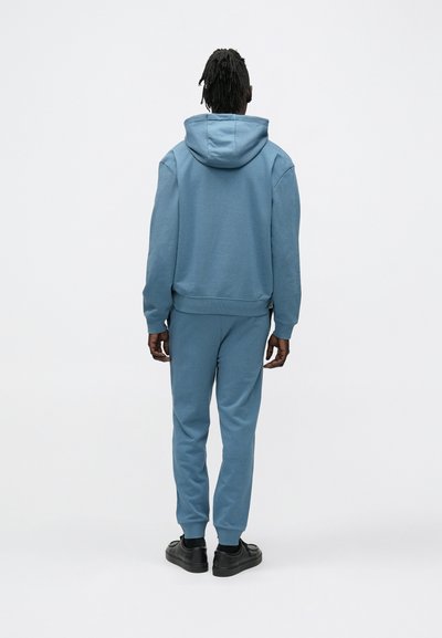 Sweat-shirt bleu clair et pantalon de jogging assorti en matière douce. Comprend une capuche, des poignets côtelés et des détails de coutures subtils. Des chaussures noires complètent le look.