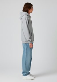 Felpa grigia con tasca a marsupio, texture morbida e cappuccio con cordino, abbinata a jeans larghi azzurri e sneakers bianche.