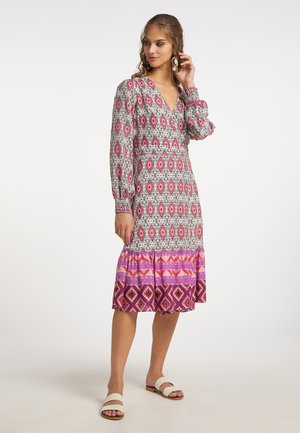 Vrouw draagt een midi-jurk met lange mouwen met rode, witte en paarse geometrische patronen, houdt één oorbel vast, staat met witte sandalen op een effen achtergrond.