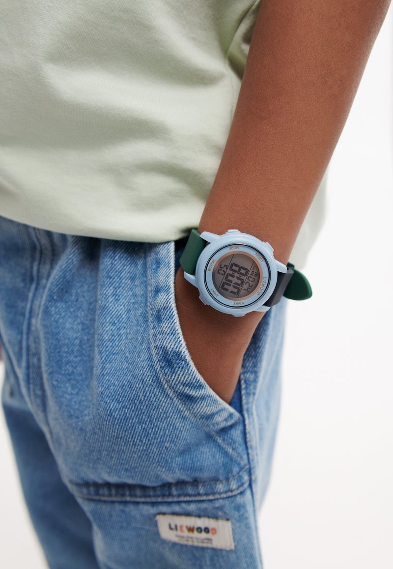 Enfant portant une montre digitale bleu clair avec un bracelet vert, main dans la poche d’un pantalon en denim bleu et une chemise de couleur claire.