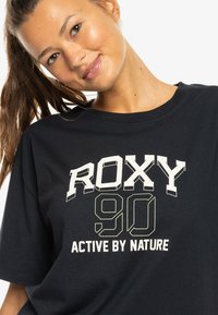 Black cotton t-shirt featuring bold white and light green text: "ROXY 90 ACTIVE BY NATURE." Crew neck and relaxed fit.