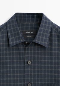 Donkerblauw geruit overhemd met een button-downkraag, twee zwarte knopen en contrasterende vierkante patronen in lichtere tinten. Merkembleem zichtbaar.