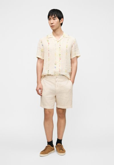 Ανοιχτό μπεζ πουκάμισο με κοντά μανίκια και πολύχρωμη κεντητή λουλουδένια διακόσμηση, συνδυασμένο με λινά σορτς στο ίδιο χρώμα, καφέ σουέντ αθλητικά παπούτσια και μαύρες κάλτσες.