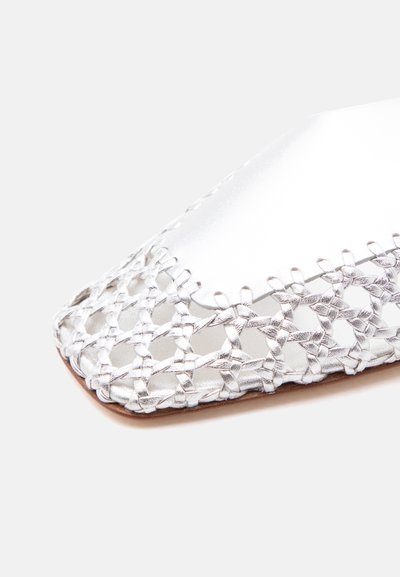 Κοντινό πλάνο στην μύτη ενός ασημένιου πλεκτού δερμάτινου παπουτσιού με ανοιχτό σχέδιο πλέξης και επίπεδη καφέ σόλα σε λευκό φόντο.