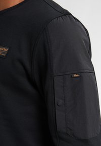 Zwarte lange mouwen shirt met textuurstof op de mouwen, voorzien van een borstpatch en details met knoopzak. Het materiaal is zacht en duurzaam.