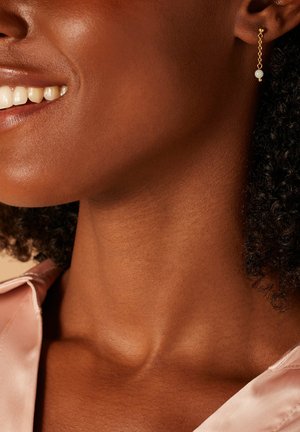Gouden kettingoorbellen met een kleine witte parel. De achtergrond toont een zachte huid en een roze satijnen blouse.