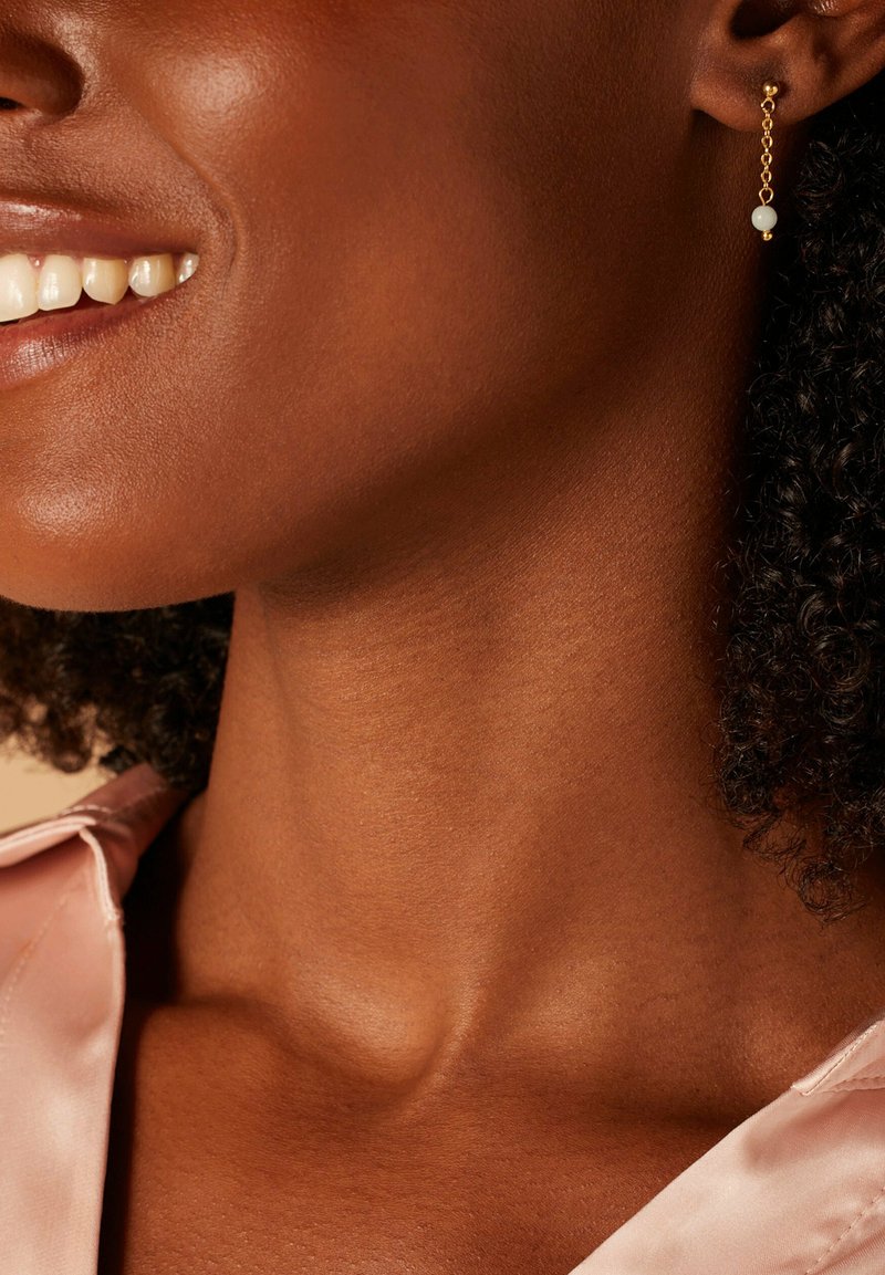Boucles d'oreilles en chaîne en ton or avec une petite perle blanche. L'arrière-plan met en valeur une peau lisse et un chemisier en satin rose.