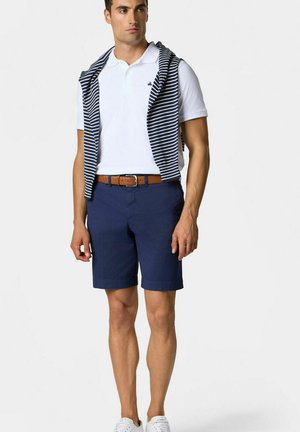Marineblauwe shorts met een gestructureerde riem, gecombineerd met een witte polo. Een gestreept navy en witte lichtgewicht trui is over de schouders gedrapeerd.