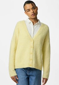 Gul, fluffig cardigan med djup V-ringning, ribbade ärmslut och sex stora knappar. Buren över en vit skjorta, tillsammans med blå jeans.