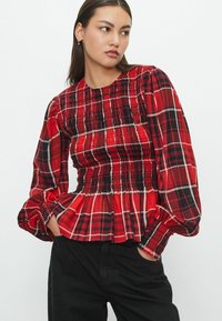 Blouse à carreaux rouge et noir avec un corsage smocké, des manches longues bouffantes et un ourlet évasé. Présente un tissu en coton texturé et des détails froncés.