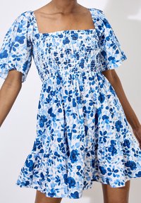 Robe fleurie avec un décolleté carré et des manches bouffantes courtes. Tissu blanc avec des motifs de fleurs bleues. Jupe évasée avec ourlet à volants.