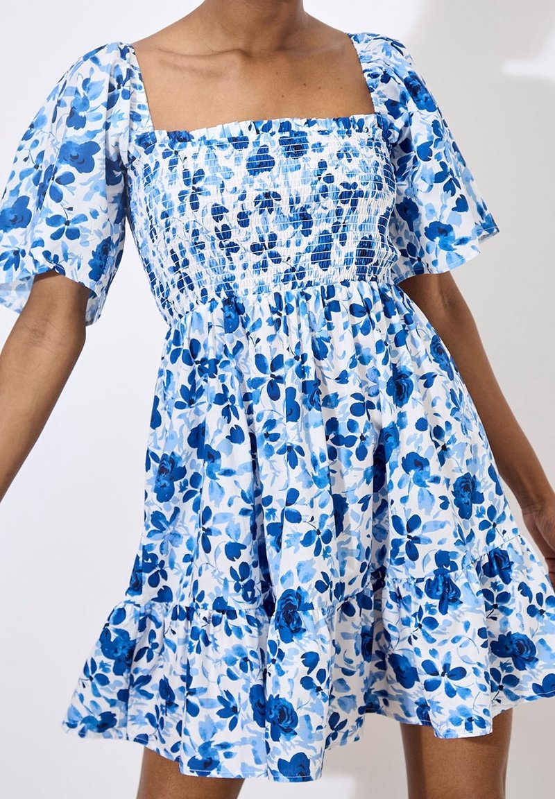 Robe fleurie avec un décolleté carré et des manches bouffantes courtes. Tissu blanc avec des motifs de fleurs bleues. Jupe évasée avec ourlet à volants.