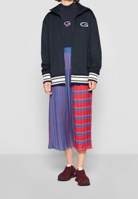 Marinblå zip-up jacka med vita ränder, svart långärmad topp, flerfärgad plisserad kjol med röda, blå och gyllene accenter, samt vinröda sneakers.