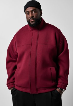 Ein Mann trägt eine bordeauxrote Jacke mit Reißverschluss, eine schwarze Mütze, schwarze Hose und eine Armbanduhr und steht vor einem schlichten grauen Hintergrund.