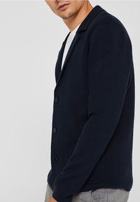 Navy knitted cardigan with a notched collar, two front pockets, and three black buttons. Fitted silhouette with ribbed texture.