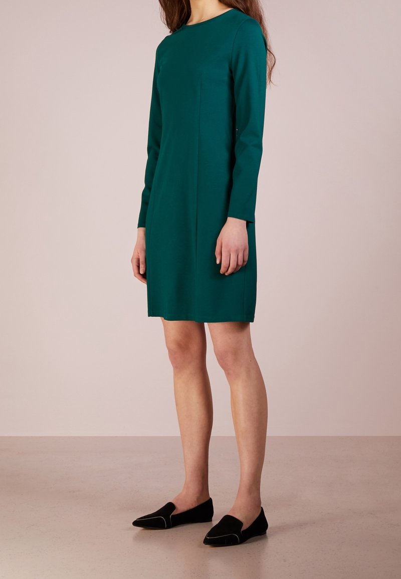 Femme portant une robe teal à manches longues, longueur genou, et des mocassins noirs, se tenant devant un fond beige uni.