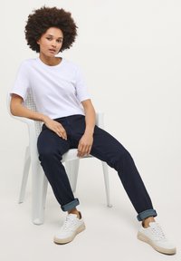 Balta īsām piedurknēm t-krekls, tumši zilas sarullētas džinses un baltas kedas ar krēmkrāsas akcentiem. Modelis sēž uz baltas plastmasas krēsla.