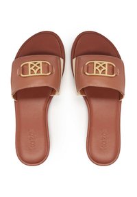 Smeđe natikače s širokim remenom koji ima zlatni logo. Potplati su ravni i glatki, izrađeni od fleksibilnog materijala.