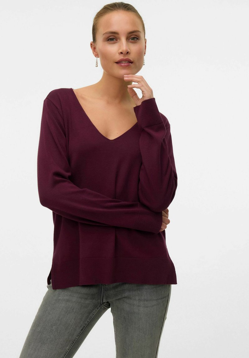 Maglione bordeaux a scollo a V con maniche lunghe, polsini e orlo a costine, abbinato a jeans grigi. Tessuto liscio con spacchi laterali per il comfort.