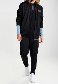 Schwarzer Sportanzug mit Reißverschlussjacke, langen Ärmeln in Hellblau und schwarzen Hosen. Verfügt über das weiße Puma-Logo. Sneakers sind nicht detailliert.