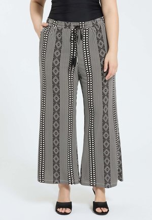 Vrouw die zwart-witte, geometrisch patroon wijde broek draagt met een trekkoord in de taille en zwarte open-toe hakken, staand met handen dicht bij de zakken.