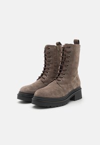 Bottines en daim marron avec un laçage à l'avant, fermeture éclair sur le côté, semelle en caoutchouc noir épais et tirant à l'arrière. Finition texturée sur l'ensemble.