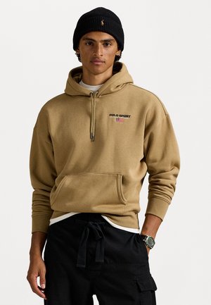 Khaki hoodie met logo en details van de Amerikaanse vlag, verstelbare trekkoord, grote voorkantzak, gedragen over zwarte broek, gecombineerd met een beanie.
