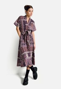 Fialové vzorované šaty s krátkými rukávy, s páskem v pase a splývavým střihem. Nosit se černými botami na platformě a podkolenkami nad kolena.