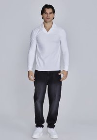 Bílá polokošile s dlouhým rukávem a límečkem, v kombinaci s tmavými džínami a bílými teniskami, stojící před neutrálním pozadím.