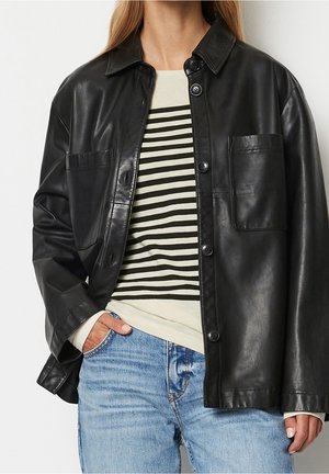 Kobieta w czarnej skórzanej kurtce zapinanej na guziki, na kremowo-czarnej pasiastej koszuli, z jasnoniebieskimi dżinsami, ręka w kieszeni.