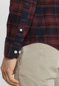 Rot-navy karierte Hemd mit langen Ärmeln, ausgestattet mit weißen Knöpfen und einem geknöpften Bündchen. Der Stoff hat eine weiche Textur, kombiniert mit beigen Hosen.