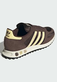 Καφέ σουέντ αθλητικό παπούτσι με πάνελ από δίχτυ, τρεις λευκές ρίγες και κίτρινες λεπτομέρειες. Διαθέτει ελαφριά σόλα με πολύχρωμες ενθέσεις για απορρόφηση κραδασμών.