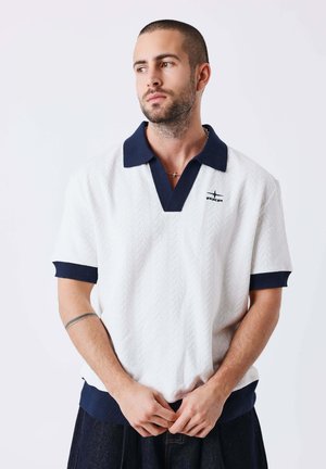 Man met kort haar en baard, gekleed in een wit shirt met korte mouwen met een marineblauwe kraag en manchetten, staand tegen een effen witte achtergrond.