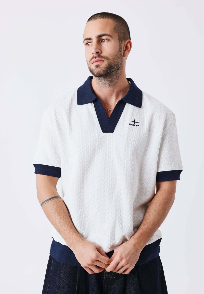Homme aux cheveux courts et à la barbe, portant une chemise blanche à manches courtes avec col et poignets bleu marine, debout devant un fond blanc uni.