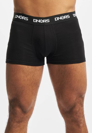 Mees, kes kannab musti bokserlühikuid, millel on bränditud vöö "DNGRS", nähtav vööst kuni reie keskosani valgel taustal.