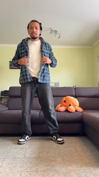 Férfi kékkockás inget visel fehér póló felett, sötét farmernadrággal és fekete-fehér tornacipővel; háttérben egy plüss narancssárga polip.