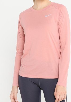 Kvinde iført en langærmet pink atletisk trøje med et lille sølv Nike-logo og mørkegrå leggings.