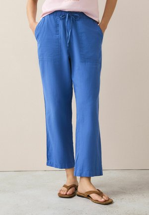 Person wearing blue drawstring wide-leg pants with front pockets and brown flip-flop sandals, standing on a light floor against a beige wall.