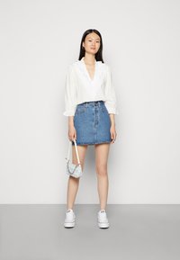 Blusa branca com decote em V em renda, mangas bufantes, combinada com uma saia mini de ganga azul e tênis brancos. Segurando uma pequena bolsa estampada.