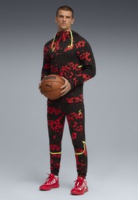 Male model in a black tracksuit with red and yellow patterns, holding a basketball, wearing red shoes, and standing against a gray background.