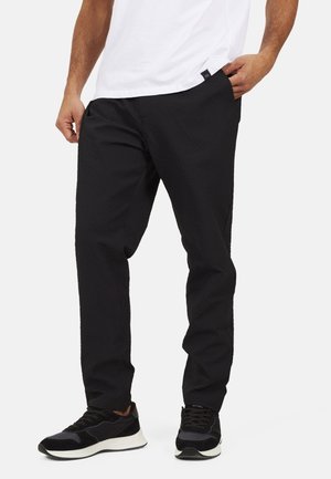 Uomo che indossa pantaloni neri testurizzati, sneaker nere con suole bianche e una maglietta bianca infilata, in piedi con le mani vicino alle tasche.