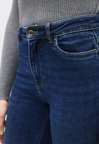 Σκούρο μπλε τζιν μεσαίας μέσης, με μεταλλικά χρυσά κουμπιά, λεπτομέρειες ξεθωριάσματος και κλασικό σχέδιο με πέντε τσέπες.