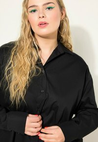 Sort skjorte med knapper i et glat stof med krave, der har lange ærmer og en afslappet pasform; hænder der justerer knapperne.