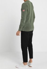 Man in een olive groene geribbelde sweater met een logo patch op de mouw, zwarte broek en witte sneakers, staande tegen een witte achtergrond.