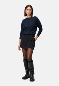 Marineblauer Strickpullover mit langen Ärmeln, schwarzer Wickelrock mit Knopfdetails, schwarze Strumpfhosen und knielange schwarze Stiefel mit einer robusten Sohle.