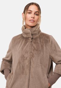 Manteau en fausse fourrure beige avec un col montant, des manches longues et des poches latérales. Présente une texture douce et un léger éclat. Idéal pour les temps frais.