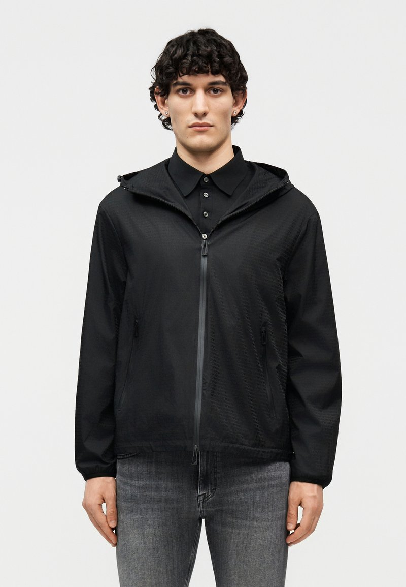 Giovane uomo con capelli ricci e scuri che indossa una giacca nera con cappuccio e zip sopra una camicia nera con colletto e jeans grigi, in piedi rivolto verso l'obiettivo.